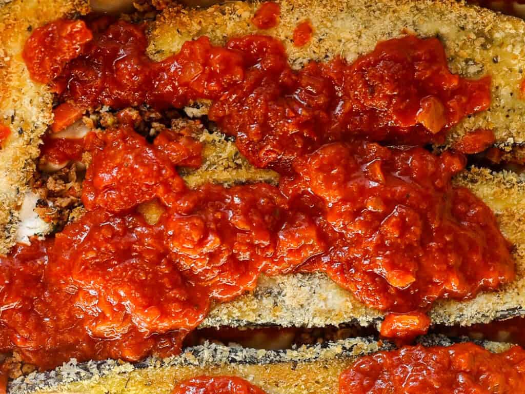 Close-up of breaded eggplant slices topped with a chunky red tomato sauce, giving a textured and savory appearance. The dish is arranged in layers, showcasing a rich and hearty presentation.